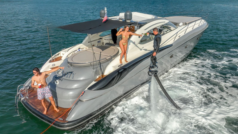 Aerial view of a luxury gray-and-white motor yacht in coastal waters during a sunny boat party, with a flyboard rider spraying water beside the stern, a woman in a bikini handing a drink from the deck, and a man holding a cup on the swim platform under an American flag.