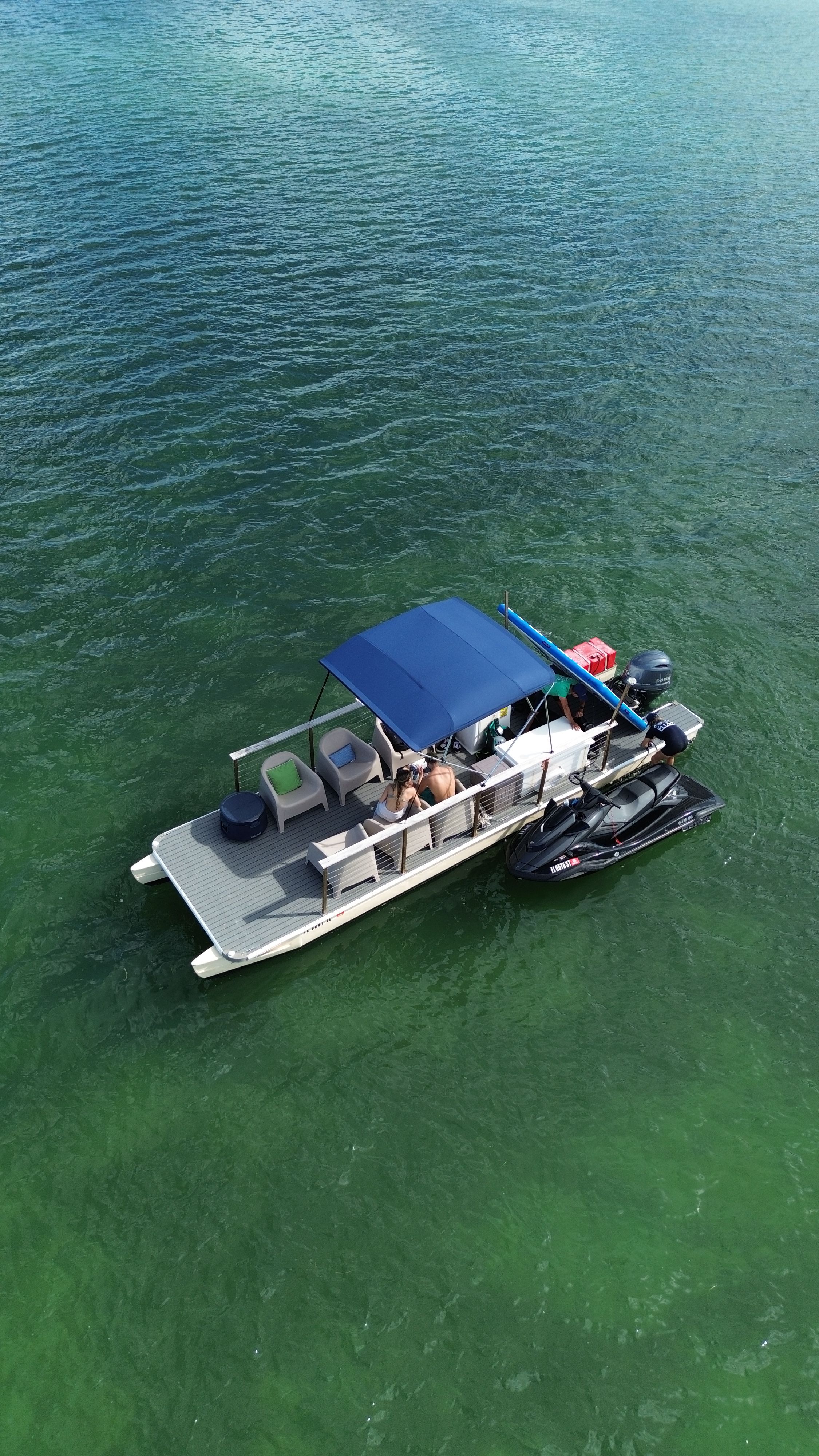 Aerial view of a pontoon boat with a blue canopy and attached black jet ski floating on clear green-turquoise water, people relaxing on the deck