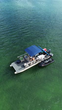 Aerial view of a pontoon boat with a blue canopy and attached black jet ski floating on clear green-turquoise water, people relaxing on the deck