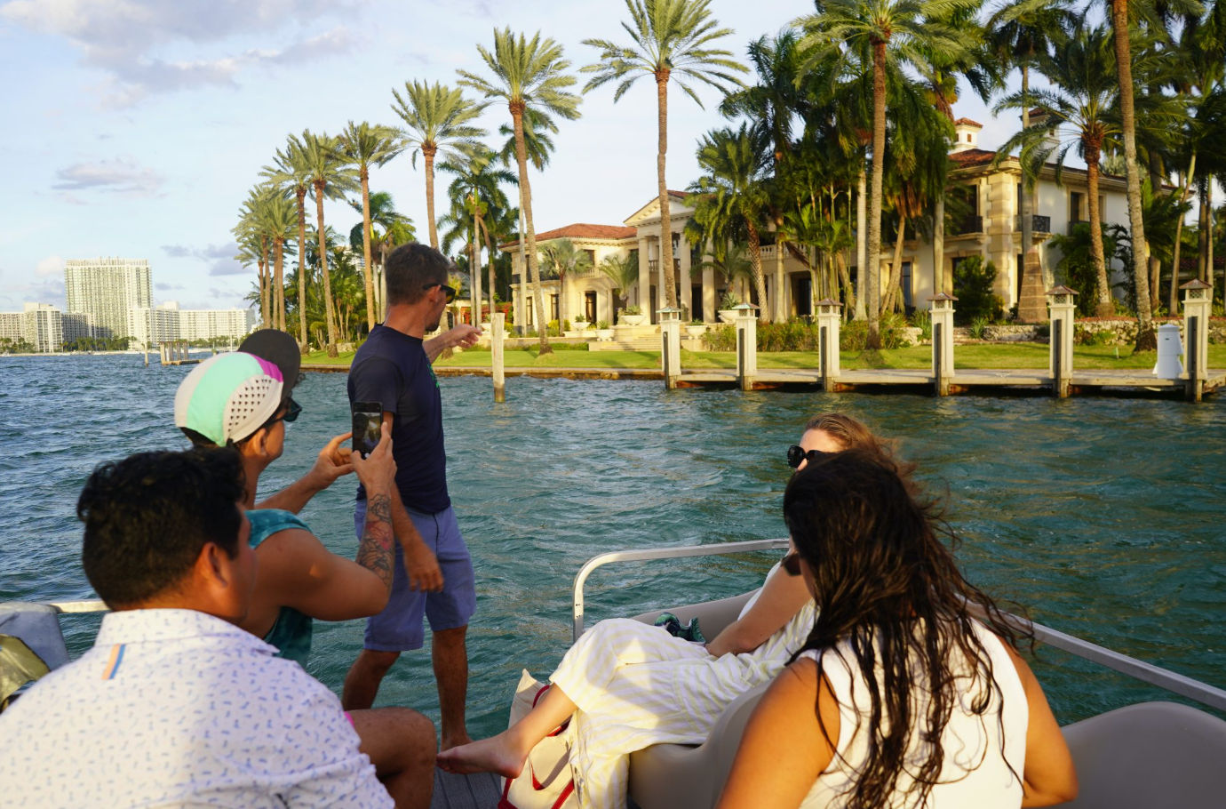 Group on a boat admiring luxury waterfront mansions and tall palm trees along the Miami shoreline on a sunny day.