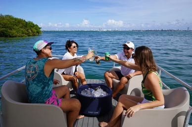 Four friends toasting drinks on a pontoon boat in a sunny tropical bay with mangroves nearby and a city skyline on the horizon