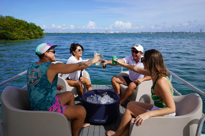 Four friends toasting drinks on a pontoon boat in a sunny tropical bay with mangroves nearby and a city skyline on the horizon