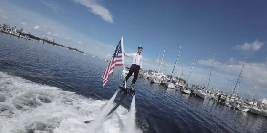Person on a flyboard holding an American flag above a coastal marina with sailboats and blue sky, high-energy waterfront water-sports scene