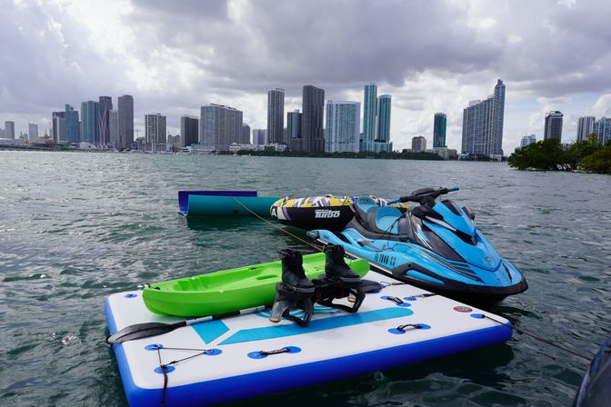 Blue jet ski and inflatable towable tied to a floating board with a green kayak and wakeboard boots on a bay, cloudy waterfront city skyline in the background.