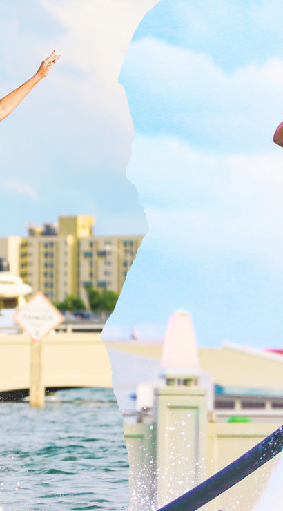 Two people flyboarding above marina water, wearing blue helmets and life vests, smiling and waving near waterfront buildings and a palm tree – coastal water sports scene.