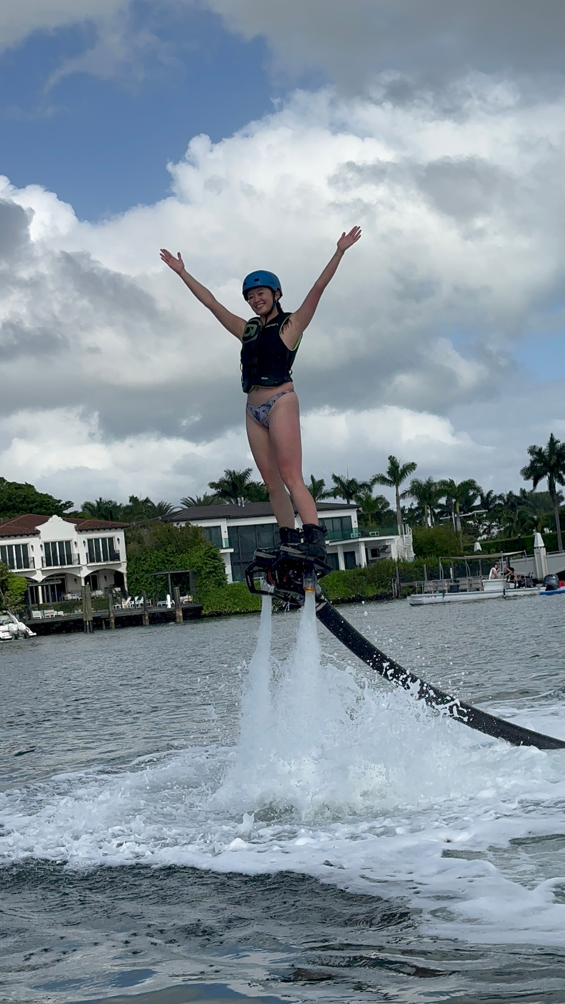 Person wearing helmet and life vest flyboarding over a tropical waterfront bay, arms raised as twin water jets lift them past palm trees and luxury homes under a cloudy blue sky