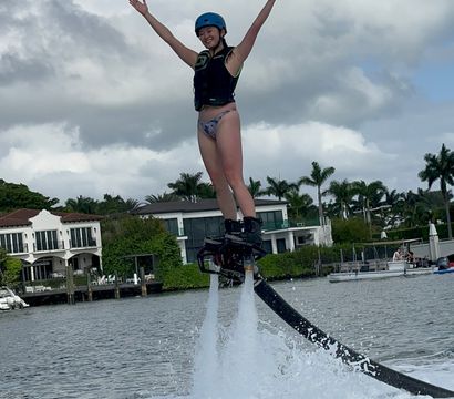 Person wearing helmet and life vest flyboarding over a tropical waterfront bay, arms raised as twin water jets lift them past palm trees and luxury homes under a cloudy blue sky