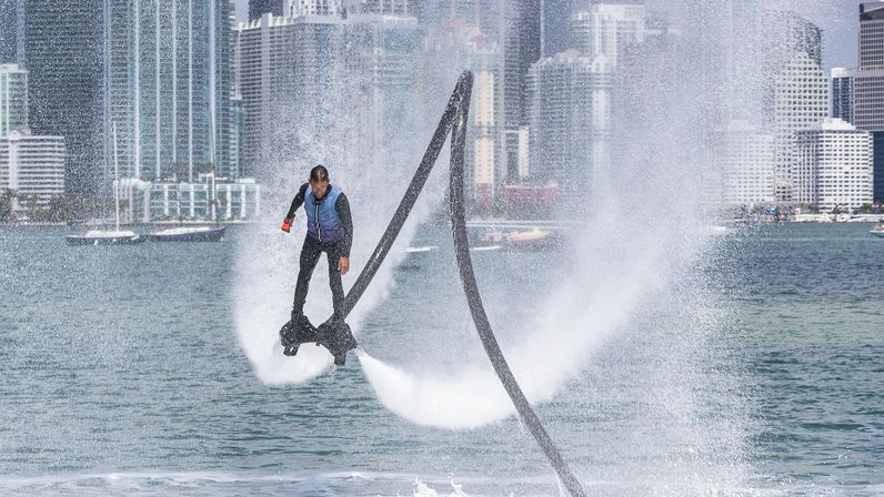 Thrilling flyboarder launching above a bay, water-jet arcs spraying around them with a downtown waterfront skyline in the background — dynamic urban water-sport scene.