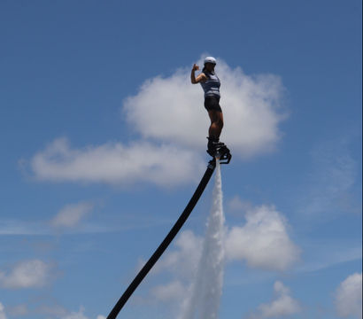 Flyboarding rider in helmet and life vest gives a thumbs-up as powerful water jets propel them high above coastal water against a bright blue sky with fluffy clouds.