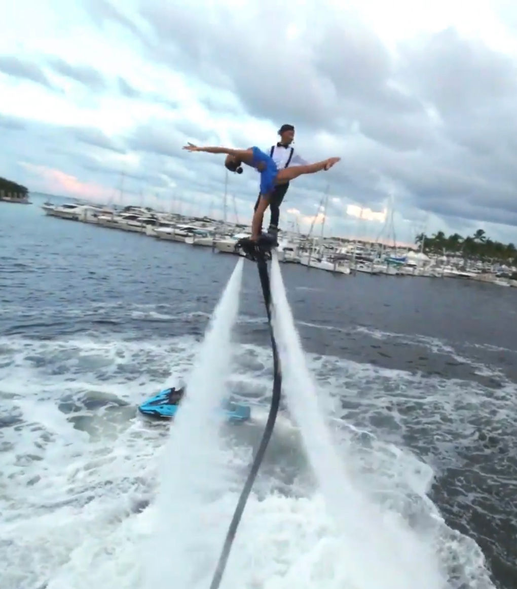 Two acrobats on a flyboard striking a dramatic split above churning water, propelled by twin water jets near a yacht-filled marina and a nearby jet ski.