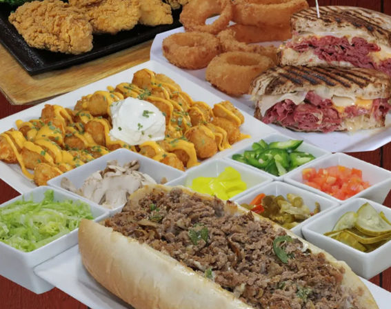 Diner-style American comfort food platter with a loaded cheesesteak sub, grilled deli sandwich, crispy onion rings, fried chicken, cheesy loaded tater tots and assorted pickles and veggies