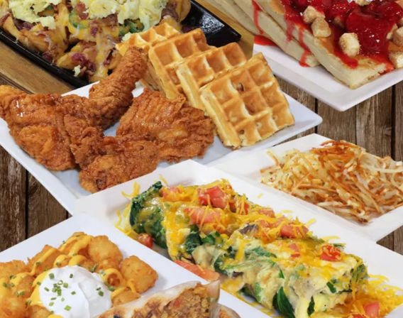 Colorful American brunch spread on a wooden table: Southern-style crispy fried chicken with golden waffles, cheesy veggie omelette, hash browns and tater tots topped with sour cream, plus a strawberry-topped waffle