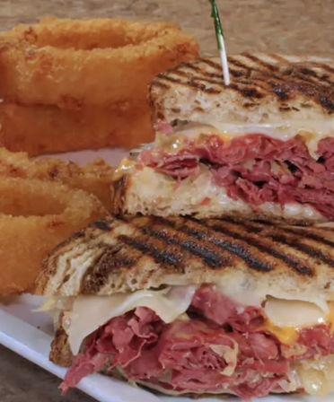 Grilled rye deli sandwich piled with thinly sliced pastrami and melted Swiss cheese, cut in half with a toothpick and served with crispy golden onion rings