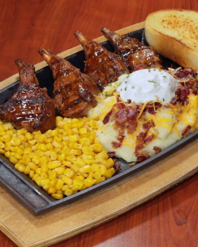 Sizzling cast-iron platter with four glazed grilled lamb chops, buttered sweet corn, loaded mashed potatoes topped with sour cream and bacon bits, and toasted garlic bread on a wooden serving board.