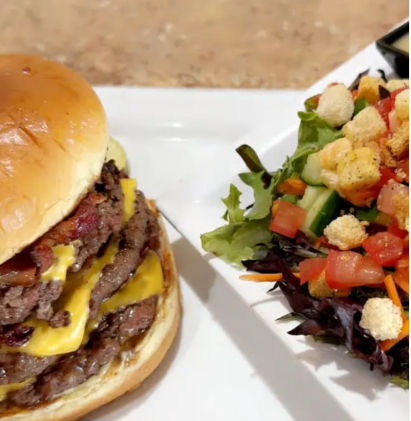 Juicy double cheeseburger with melted cheddar on a brioche bun next to a fresh mixed green salad with tomatoes, cucumber, shredded carrots and croutons on a white plate.