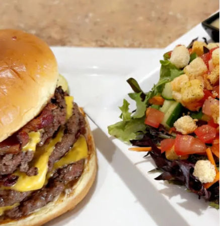 Juicy double cheeseburger with melted cheddar on a brioche bun next to a fresh mixed green salad with tomatoes, cucumber, shredded carrots and croutons on a white plate.