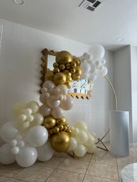 Gold and white balloon arch on a gold hoop stand in a modern indoor entryway, metallic gold, cream and white balloon clusters layered around a mirror backdrop and tile floor