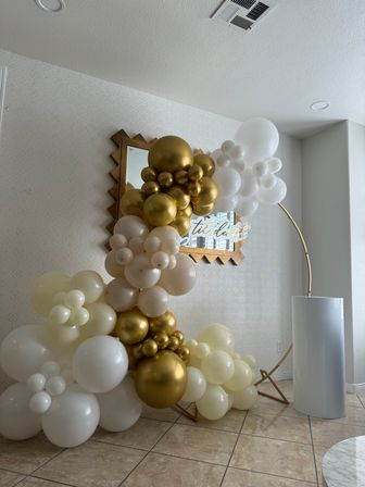 Gold and white balloon arch on a gold hoop stand in a modern indoor entryway, metallic gold, cream and white balloon clusters layered around a mirror backdrop and tile floor