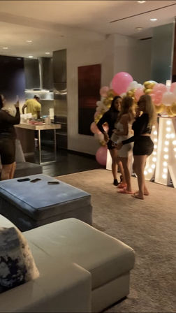 Three women pose by a pink, white and gold balloon arch and illuminated marquee letters in a modern apartment living room during a celebration, while a guest takes a photo, someone is in the kitchen, and phones rest on an ottoman.