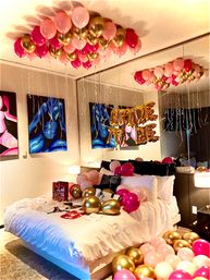 Cheerful bedroom bridal surprise with pink and gold helium balloons clustered on the ceiling and scattered across a white bed, mirrored wall reflecting gold foil 'BRIDE' letters and colorful abstract art.