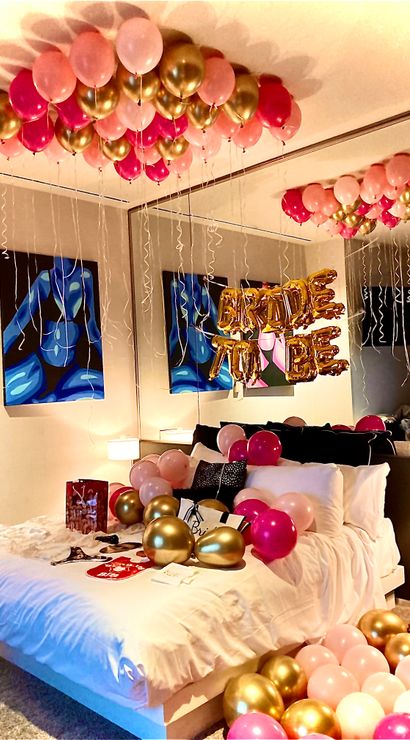 Cheerful bedroom bridal surprise with pink and gold helium balloons clustered on the ceiling and scattered across a white bed, mirrored wall reflecting gold foil 'BRIDE' letters and colorful abstract art.
