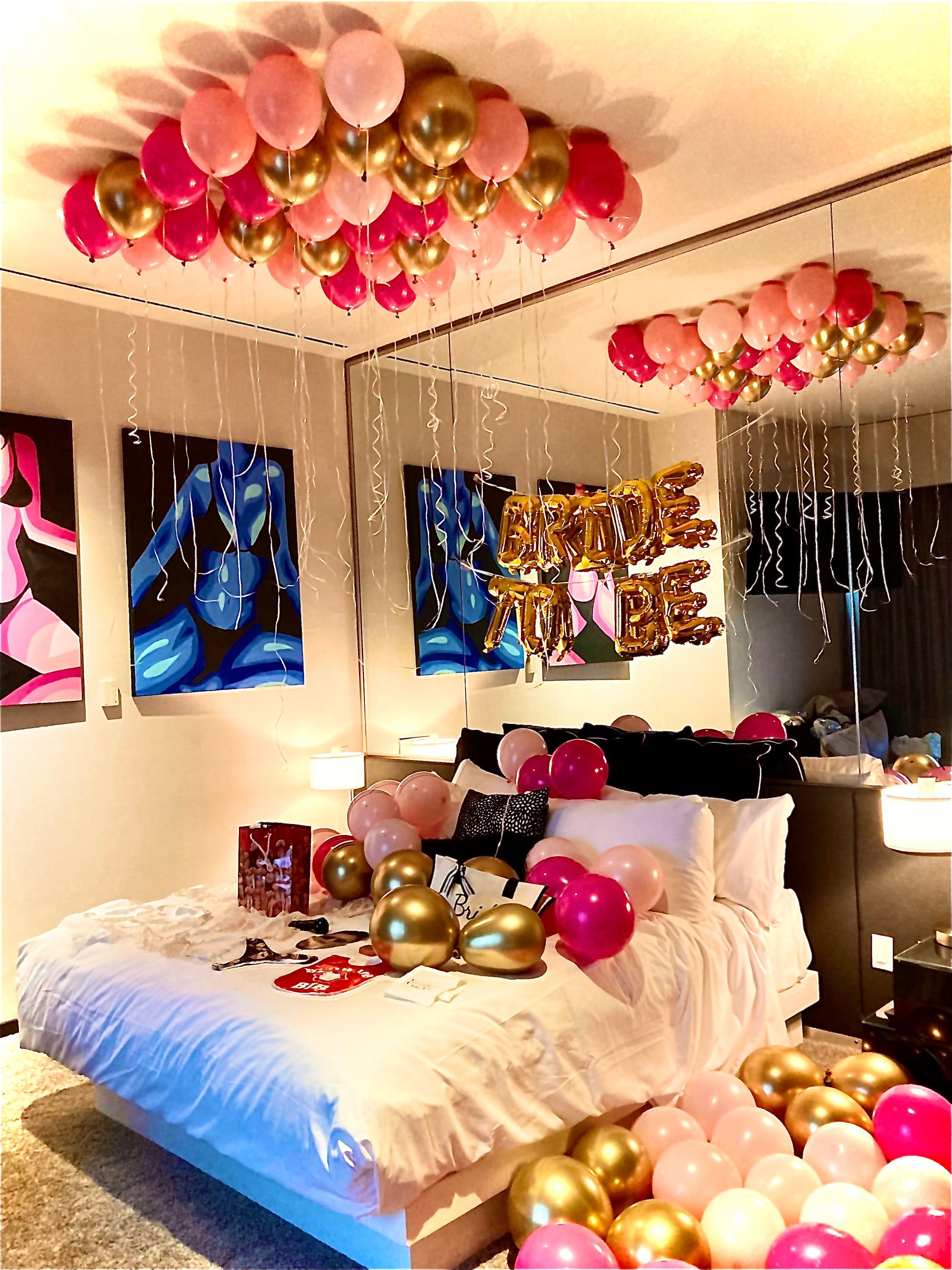 Cheerful bedroom bridal surprise with pink and gold helium balloons clustered on the ceiling and scattered across a white bed, mirrored wall reflecting gold foil 'BRIDE' letters and colorful abstract art.