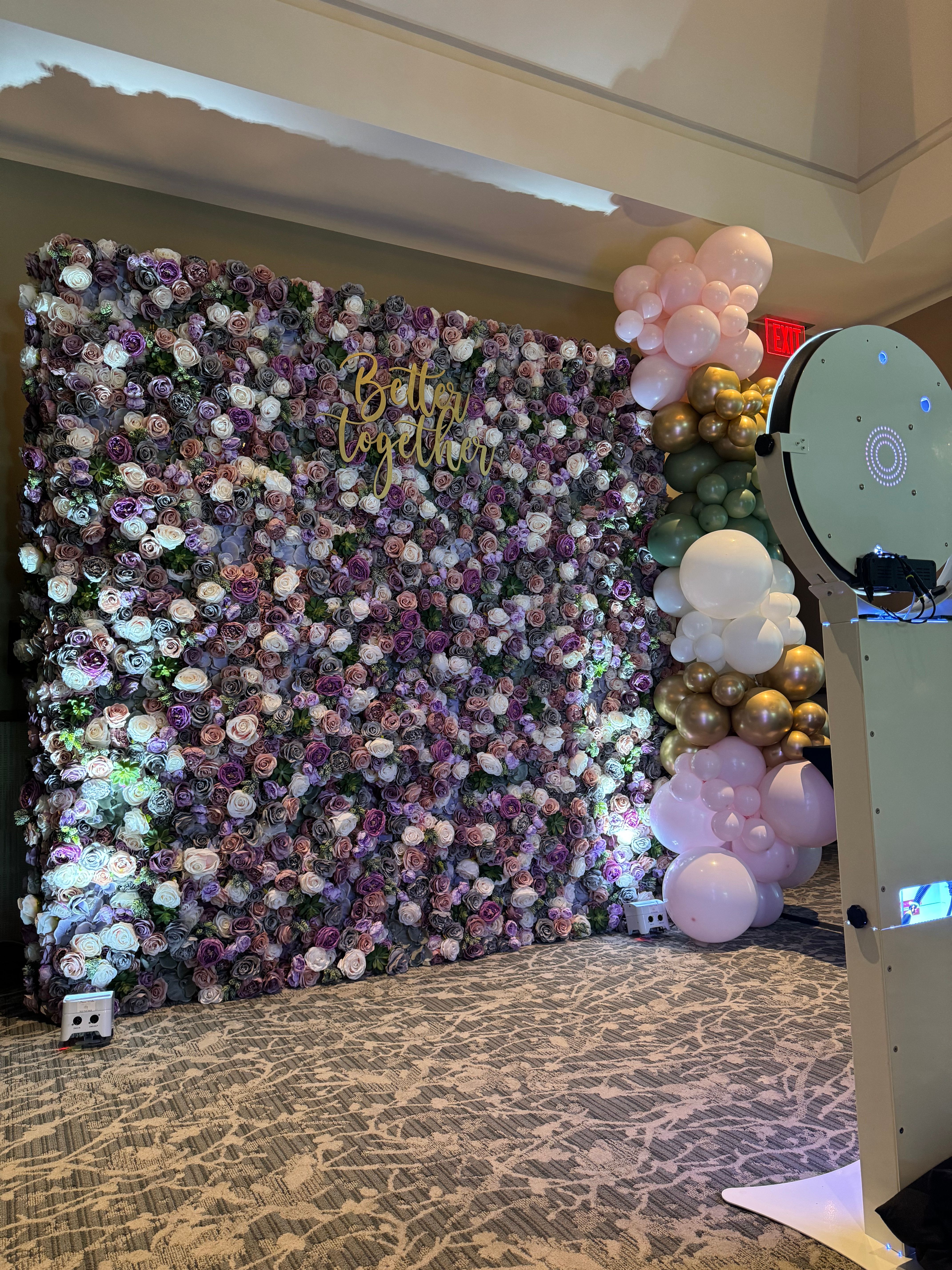 Indoor event photo backdrop with dense purple and blush rose wall, gold "Better Together" sign, pastel pink, green and gold balloon garland, and a white photo booth
