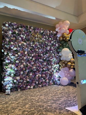 Indoor event photo backdrop with dense purple and blush rose wall, gold "Better Together" sign, pastel pink, green and gold balloon garland, and a white photo booth