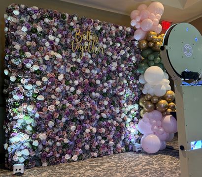 Indoor event photo backdrop with dense purple and blush rose wall, gold "Better Together" sign, pastel pink, green and gold balloon garland, and a white photo booth