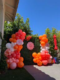 Bright backyard first-birthday setup with orange, coral, pink and white balloon garlands framing a green hedge backdrop and round birthday signs under a clear blue sky