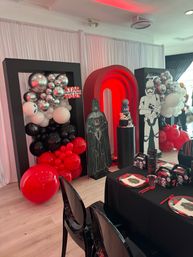 Indoor event space decorated for a Star Wars birthday party with a red LED arch, Darth Vader and stormtrooper standees, black‑red‑silver balloon clusters, a themed tiered cake on a pedestal, and a black‑draped kids table with matching plates and favor boxes.
