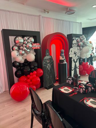 Indoor event space decorated for a Star Wars birthday party with a red LED arch, Darth Vader and stormtrooper standees, black‑red‑silver balloon clusters, a themed tiered cake on a pedestal, and a black‑draped kids table with matching plates and favor boxes.