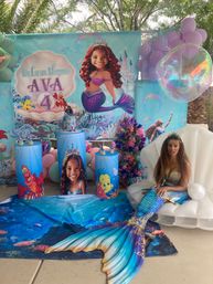 Little Mermaid–themed backyard kids' birthday setup with a large mermaid backdrop and number 4 banner, sea-creature podiums, pastel balloons, floral display, and a performer in a shimmering blue-green mermaid tail seated in a shell chair.