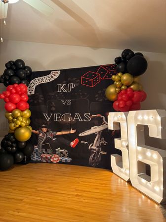 Indoor Vegas-themed 36th birthday photo backdrop with black, red and gold balloon garlands, giant illuminated marquee "36", poker chips, dice and playing-card graphics on a black backdrop over hardwood floor.