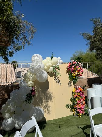 Sunny backyard backdrop with ivory and white balloon arch and vibrant pink-yellow floral garlands on a beige panel, white folding chairs under a clear blue sky.