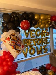 Vegas-themed hotel room balloon display with gold letter balloons reading 'VEGAS BEFORE VOWS', black, red, white and gold balloon garlands, oversized queen of hearts card and casino chip balloons by a city skyline window.