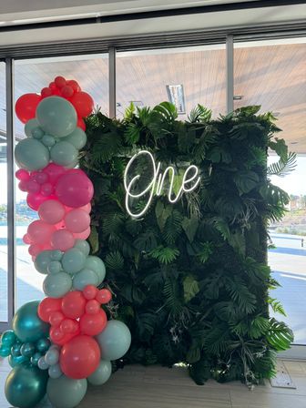 Tropical greenery wall with neon 'One' sign and teal, pink and coral balloon garland beside floor-to-ceiling windows — first birthday party backdrop.