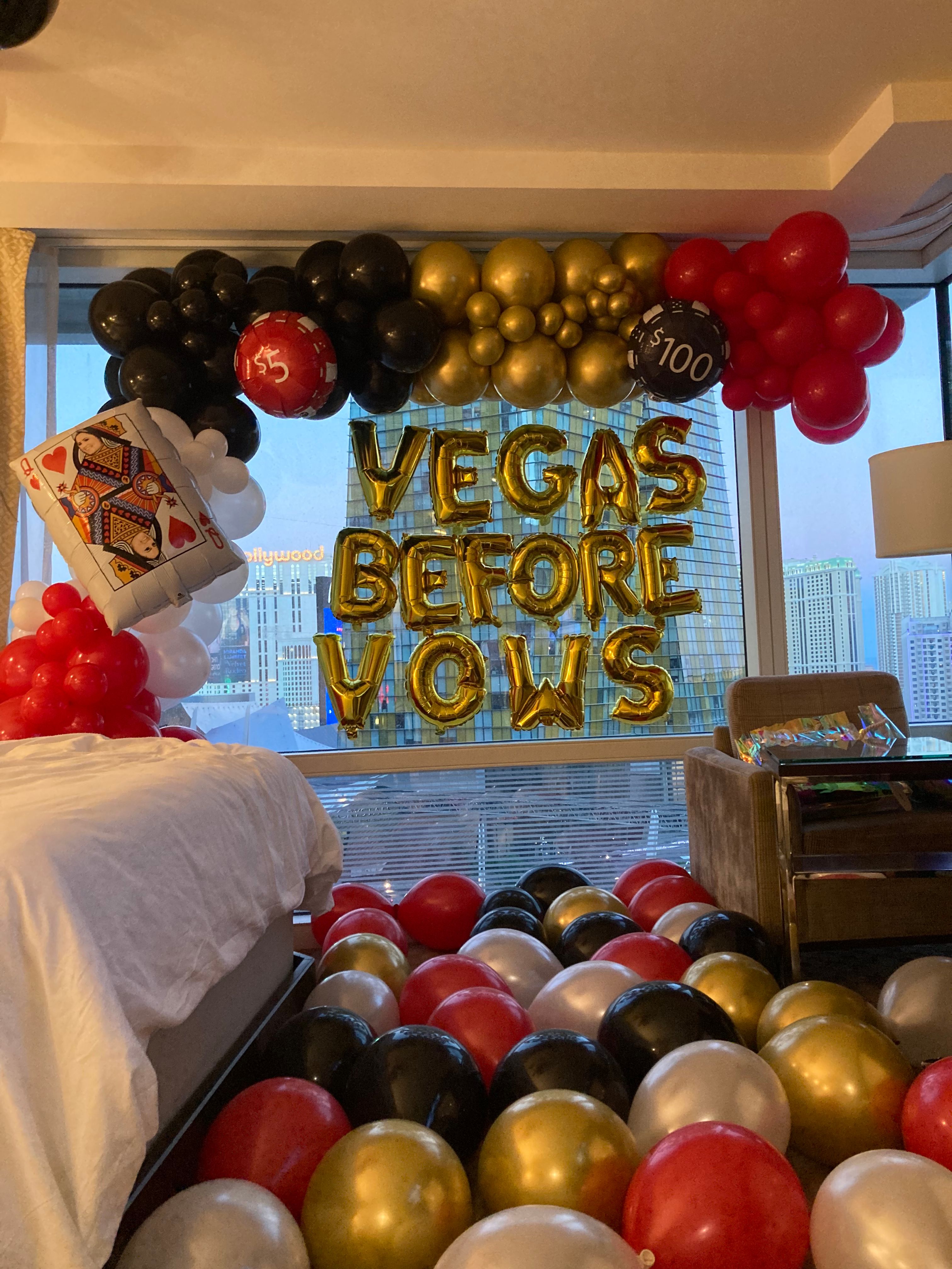 Las Vegas hotel room bachelorette display with gold foil "VEGAS BEFORE VOWS" letter balloons, red, black, gold and white balloon arch, oversized poker card and casino-chip balloons, and city skyline view