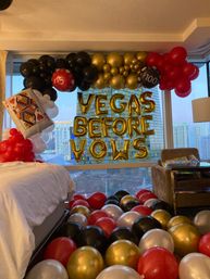 Las Vegas hotel room bachelorette display with gold foil "VEGAS BEFORE VOWS" letter balloons, red, black, gold and white balloon arch, oversized poker card and casino-chip balloons, and city skyline view