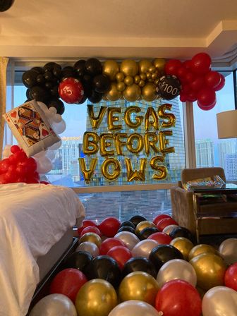 Las Vegas hotel room bachelorette display with gold foil "VEGAS BEFORE VOWS" letter balloons, red, black, gold and white balloon arch, oversized poker card and casino-chip balloons, and city skyline view