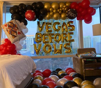 Las Vegas hotel room bachelorette display with gold foil "VEGAS BEFORE VOWS" letter balloons, red, black, gold and white balloon arch, oversized poker card and casino-chip balloons, and city skyline view