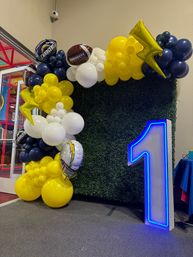Football-themed balloon arch in navy, yellow and white with team logo and football balloons, lightning-bolt accents, green hedge backdrop and illuminated blue number 1 for a first birthday party