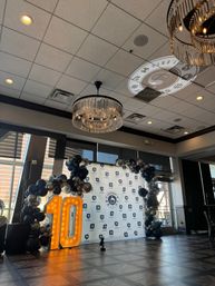 Banquet hall event setup for a 10th anniversary: illuminated marquee '10', black-and-silver balloon arch framing a branded step-and-repeat backdrop, crystal chandeliers and logo projection on the ceiling.