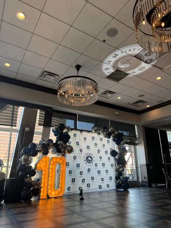 Banquet hall event setup for a 10th anniversary: illuminated marquee '10', black-and-silver balloon arch framing a branded step-and-repeat backdrop, crystal chandeliers and logo projection on the ceiling.