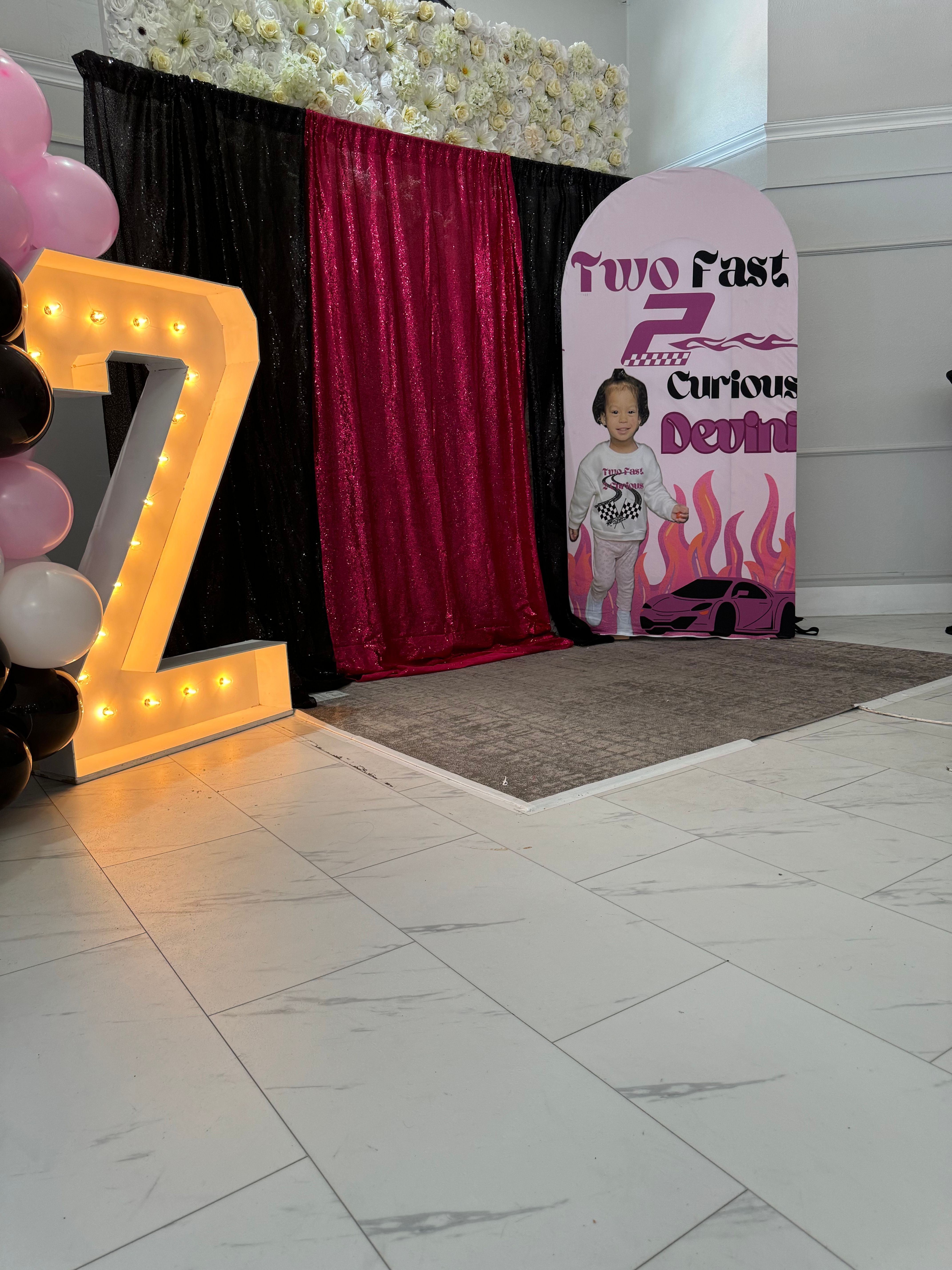 Indoor birthday photo backdrop with illuminated marquee Z, black and fuchsia sequin curtains, pink and black balloon column, floral wall and race-car themed child cutout on white tile floor