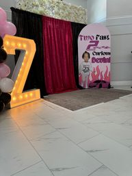Indoor birthday photo backdrop with illuminated marquee Z, black and fuchsia sequin curtains, pink and black balloon column, floral wall and race-car themed child cutout on white tile floor