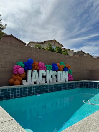 Backyard swimming pool with large white marquee letters reading JACKSON, framed by a colorful balloon garland along a beige cinderblock wall and suburban homes under a bright cloudy sky.