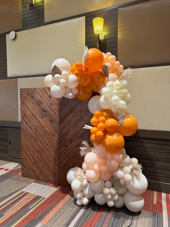 Playful orange, peach, and white balloon garland with dried palm leaves and pampas grass climbing a wooden chevron backdrop in an indoor event space under warm wall sconces.