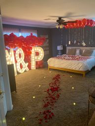 Romantic bedroom surprise with illuminated marquee letters, ceiling clusters of red balloons and hanging notes, a trail of red rose petals and LED candles leading to a petal‑covered bed.