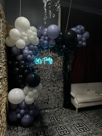 Indoor event lounge with black, white and lavender balloon arch framing a sequin backdrop, teal neon “Let’s Party” sign, hanging crystal decor and a white loveseat.